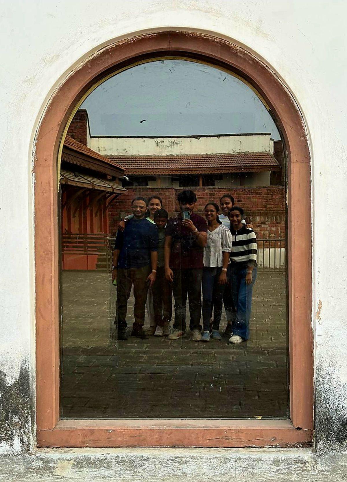 Seven of us reflected in the glass of an arched window, a tiled courtyard and brick wall behind
